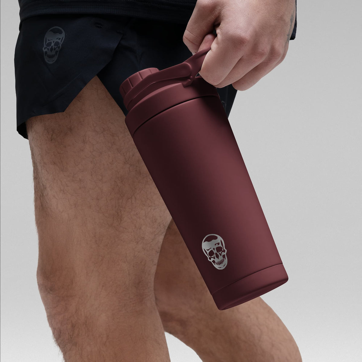 Athlete holding the Burgundy red stainless steel shaker bottle by the built in carry loop handle with the skull logo facing outward