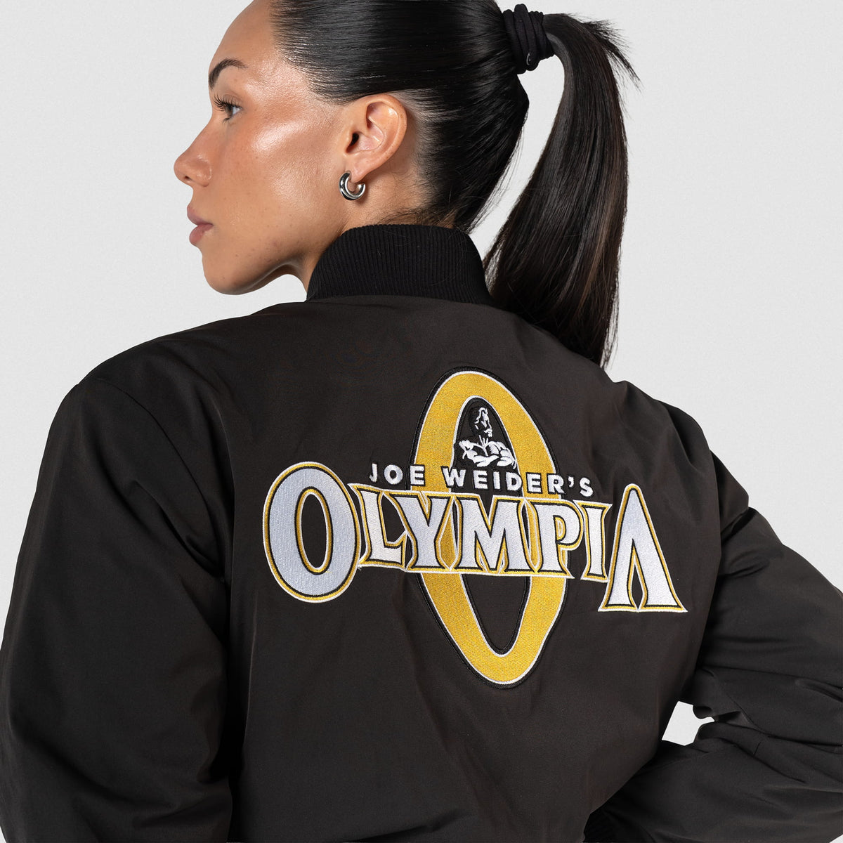 Champions Gold Crop Bomber Jacket | Close p view of the Champions Gold large back logo that says "Joe Weider's Olympia"