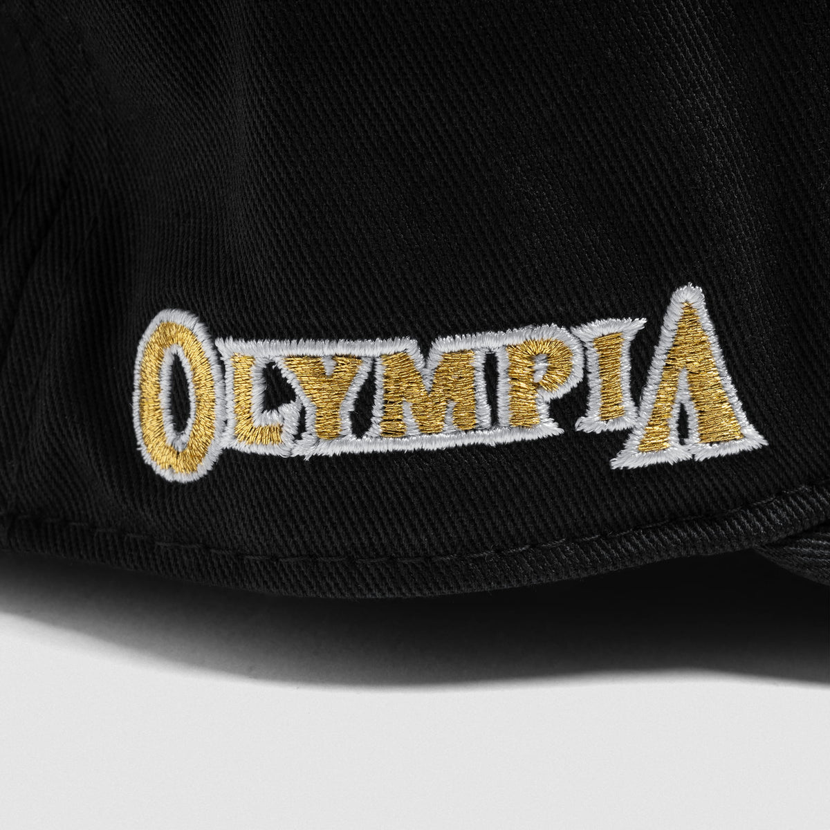 Close up of side logo stitched into the Champions Gold 5-Panel Hat that has the Olympia bodybuilding wordmark in gold text