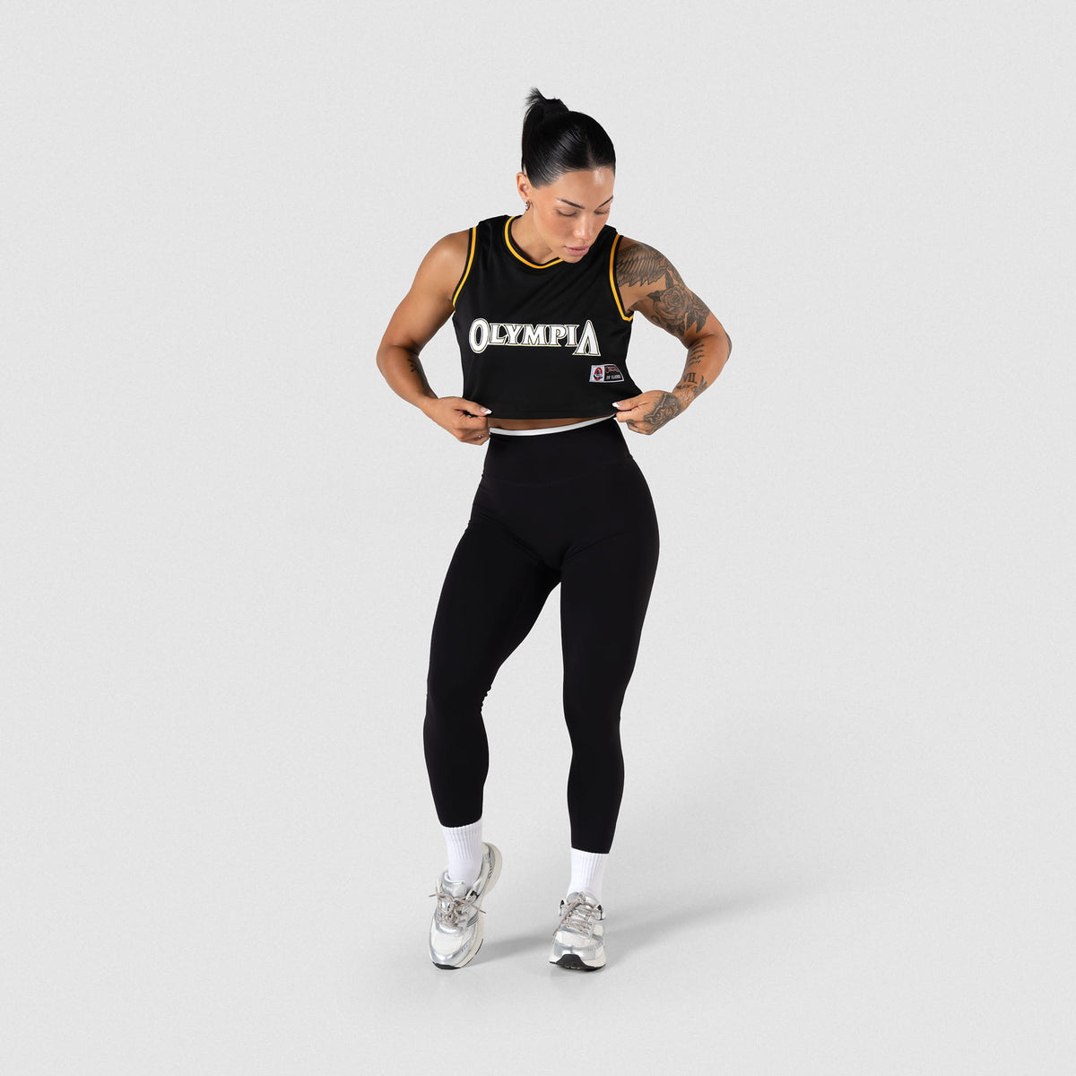 Champions Gold Crop Jersey | Model adjusting the hem of the black cropped jersey while wearing matching leggings