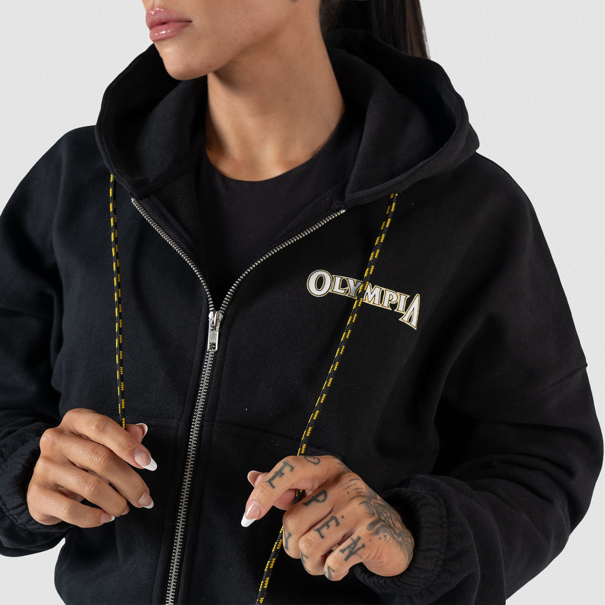 Champions Gold Cropped Zip-Up Hoodie | On model, close up of half zipped hoodie while model holds the alternating yellow and black striped hoodie drawstrings