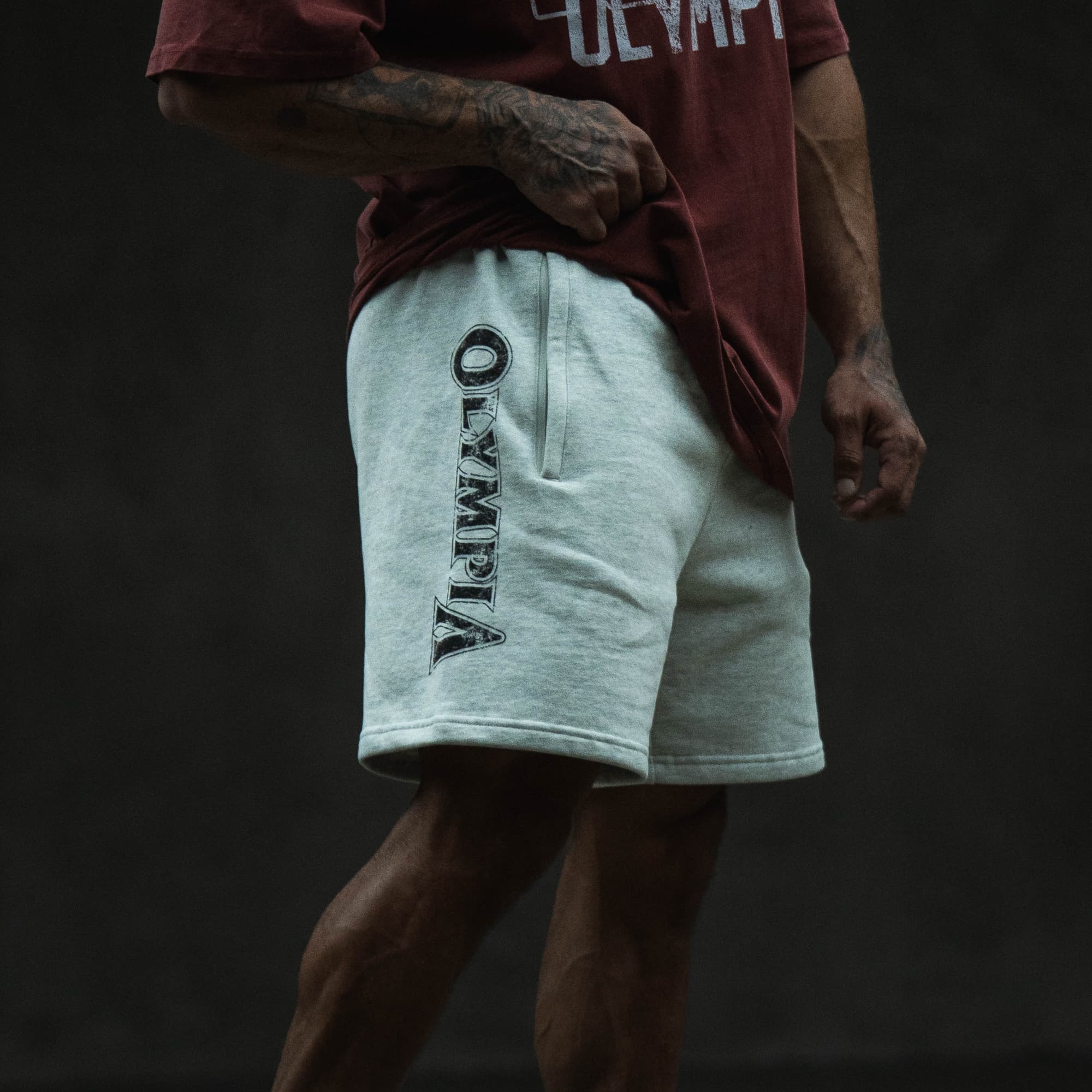 Lifestyle side view of the Olympia Sweat Short worn with a red tee, focusing on the vertical “Olympia” leg graphic.