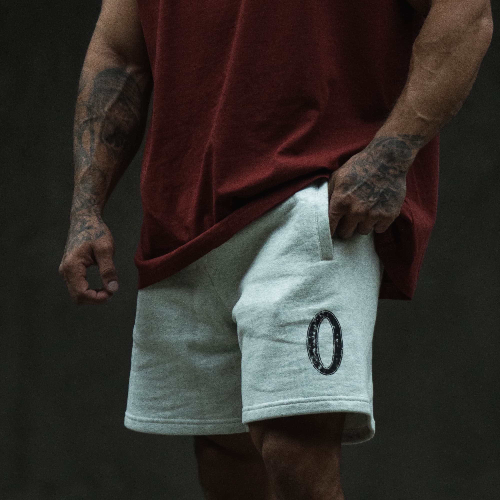 Lifestyle front view of the Olympia Sweat Short worn with a red tee, highlighting the oval wreath graphic on the left leg.