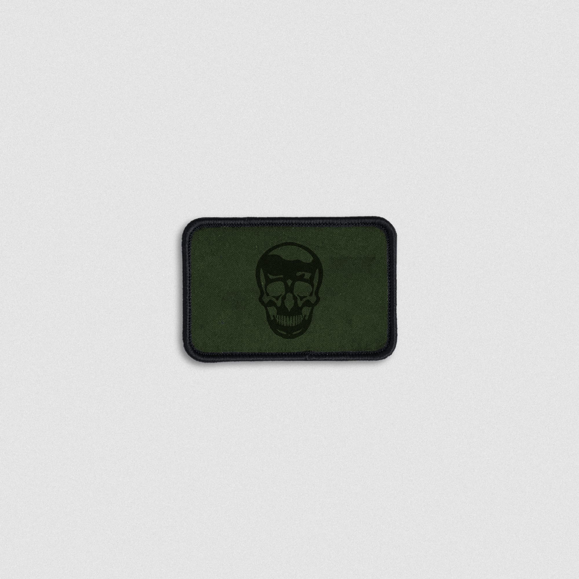 Rectangular olive green patch featuring a black skull design in the center with a black stitched border and hook-and-loop backing.